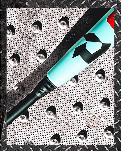 A close-up of the 2026 DeMarini The Goods (-3) BBCOR Baseball Bat (WBD2588010) rests on textured metal, showcasing its bold geometric logo and X14 Alloy Barrel as sunlight casts sharp shadows.