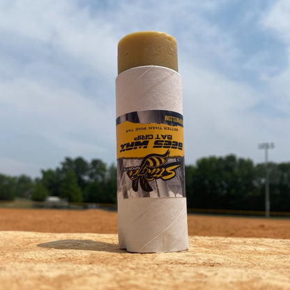 A Stinger BEES WAX Bat Grip by Stinger Sports stands upright on a baseball field, trees and blue sky behind. The label shows a bee with bold text, highlighting this natural grip as an excellent pine tar alternative.