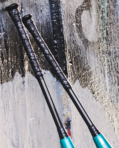 Two DeMarini 2026 Omega (-3) BBCOR baseball bats, featuring black handles and teal X14 Alloy barrels, lean against a graffiti-covered wall.