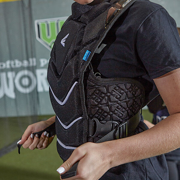 A person adjusts the straps of a black chest protector while standing in an indoor sports facility. The chest protector features textured padding and white chevron markings.