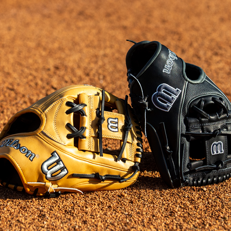 Two Wilson baseball gloves, one tan and one black, rest on a baseball field with a textured dirt surface. The gloves are positioned side by side, facing each other.