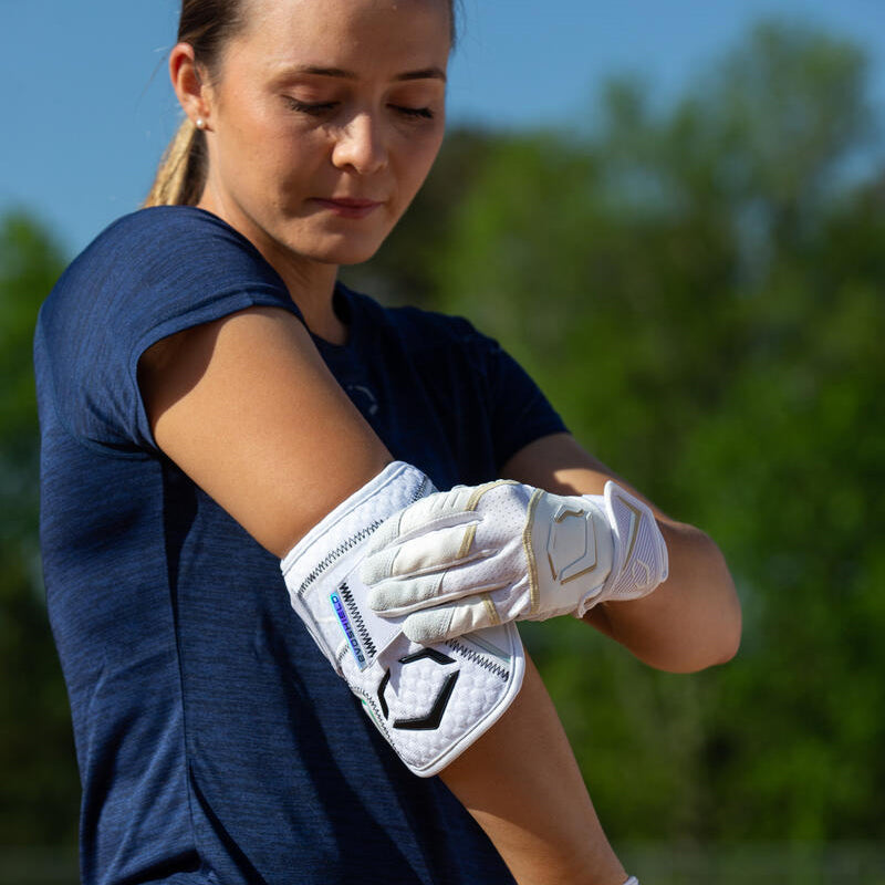 Outdoors, a woman in a navy blue shirt adjusts her EvoShield PRO-SRZ 2.0 Batter's Elbow Guard: WB572600 with Gel-to-Shell Technology, paired with white gloves. Green trees and a blue sky are visible in the background.