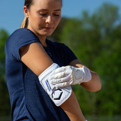 Outdoors, a woman in a navy blue shirt adjusts her EvoShield PRO-SRZ 2.0 Batter's Elbow Guard: WB572600 with Gel-to-Shell Technology, paired with white gloves. Green trees and a blue sky are visible in the background.