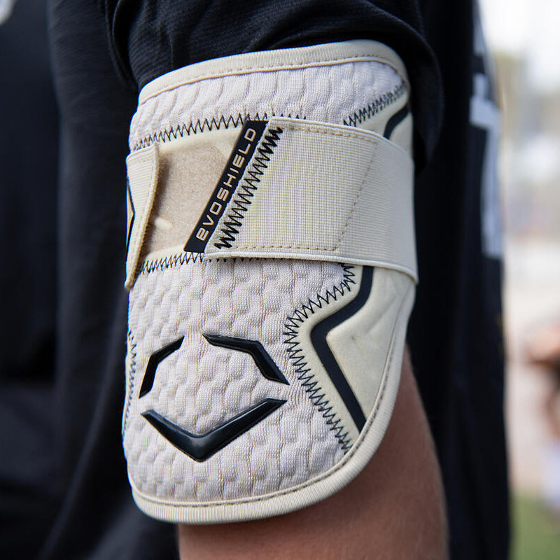 A close-up of a person's arm wearing an EvoShield PRO-SRZ 2.0 Batter's Elbow Guard (WB572600) in beige and black, with Gel-to-Shell Technology and a Velcro strap, against a blurred outdoor background.