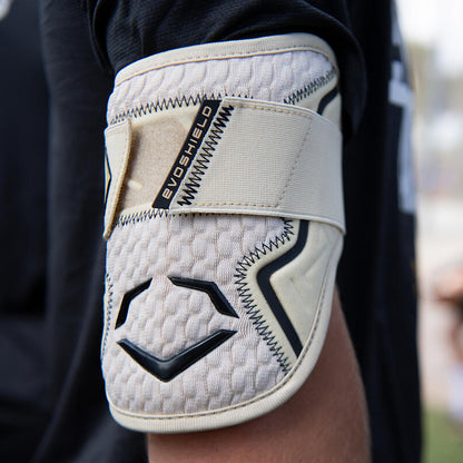A close-up of a person's arm wearing an EvoShield PRO-SRZ 2.0 Batter's Elbow Guard (WB572600) in beige and black, with Gel-to-Shell Technology and a Velcro strap, against a blurred outdoor background.