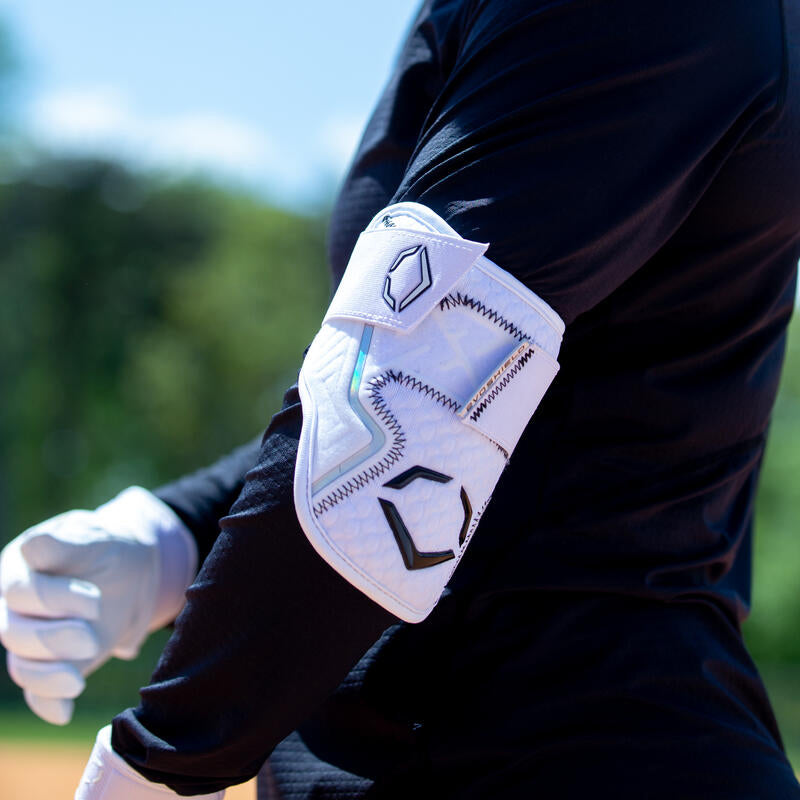 A person in a black long-sleeve shirt and white glove wears an EvoShield PRO-SRZ 2.0 Batter's Elbow Guard (WB572600), featuring black details, secured on their elbow against a backdrop of blurred greenery and blue sky.