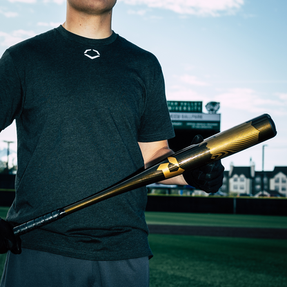 A person in a dark t-shirt and gloves stands on a baseball field, holding the 2024 DeMarini Voodoo One (-3) BBCOR Baseball Bat (WBD2461010). The background shows a scoreboard and buildings under partly cloudy skies.