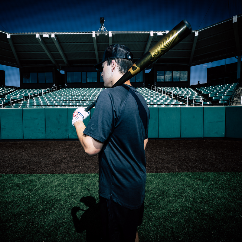 A baseball player in a black shirt and cap stands on a field, holding the 2024 DeMarini Voodoo One (-3) BBCOR Baseball Bat (WBD2461010) over his shoulder, facing empty stadium seats beneath a clear blue sky.