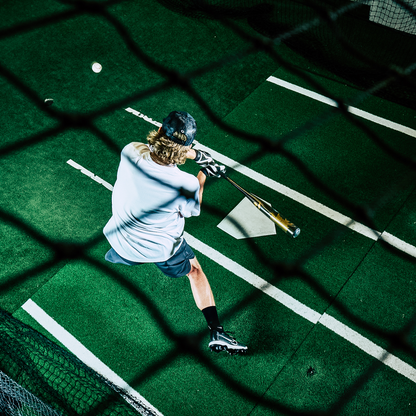 A baseball player in cleats and black shorts swings a 2024 DeMarini Voodoo One (-3) BBCOR Baseball Bat (WBD2461010) over home plate on green turf, viewed from above through netting in a batting cage.