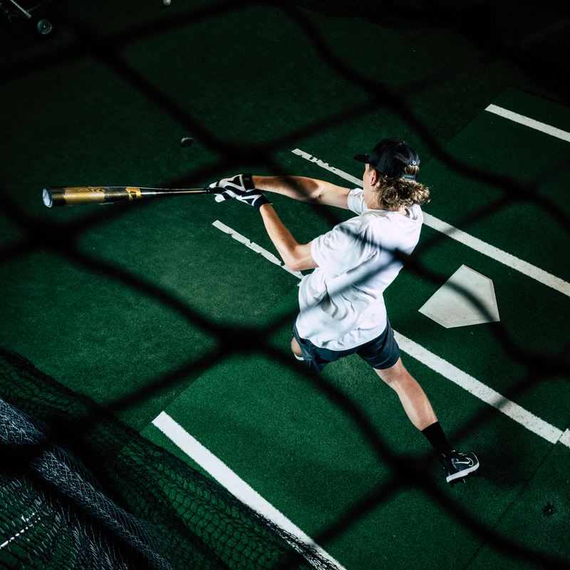 A person in a white shirt, shorts, and black cap swings the 2024 DeMarini Voodoo One (-3) BBCOR Baseball Bat (WBD2461010) in an indoor batting cage, seen from above through a net; home plate and white lines show on the green turf.