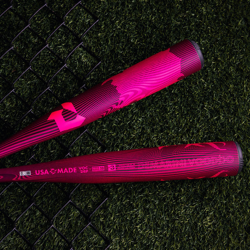 Two pink and black baseball bats with modern designs rest on artificial green turf next to a black chain-link fence. The text DeMarini Voodoo and USA Made are visible on one bat.