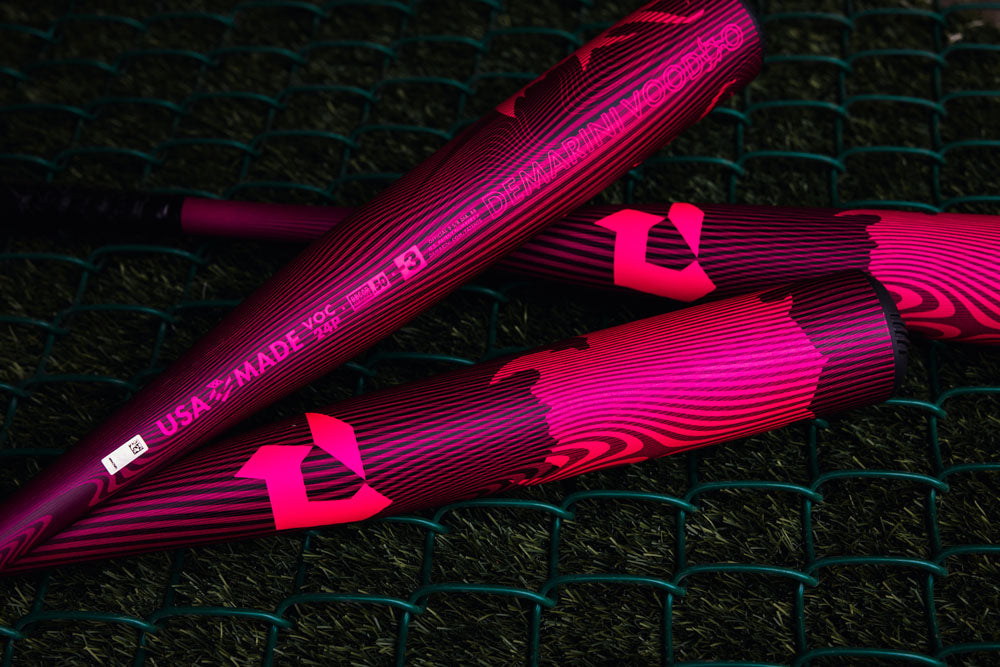 Three DeMarini 2024 Voodoo One Neon Pink (-3) BBCOR Baseball Bats (WBD2557010) with bold patterns and "USA MADE" text rest on green netting over artificial grass.