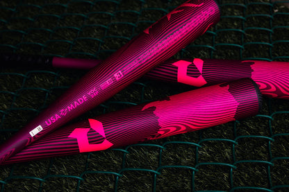 Three DeMarini 2024 Voodoo One Neon Pink (-3) BBCOR Baseball Bats (WBD2557010) with bold patterns and "USA MADE" text rest on green netting over artificial grass.