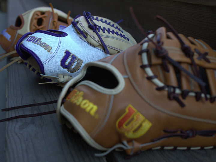 Three Wilson baseball gloves rest on wooden bleachers. One glove is tan with gold and red logo details, another is white with purple accents, and the third is tan with purple lacing and logo.