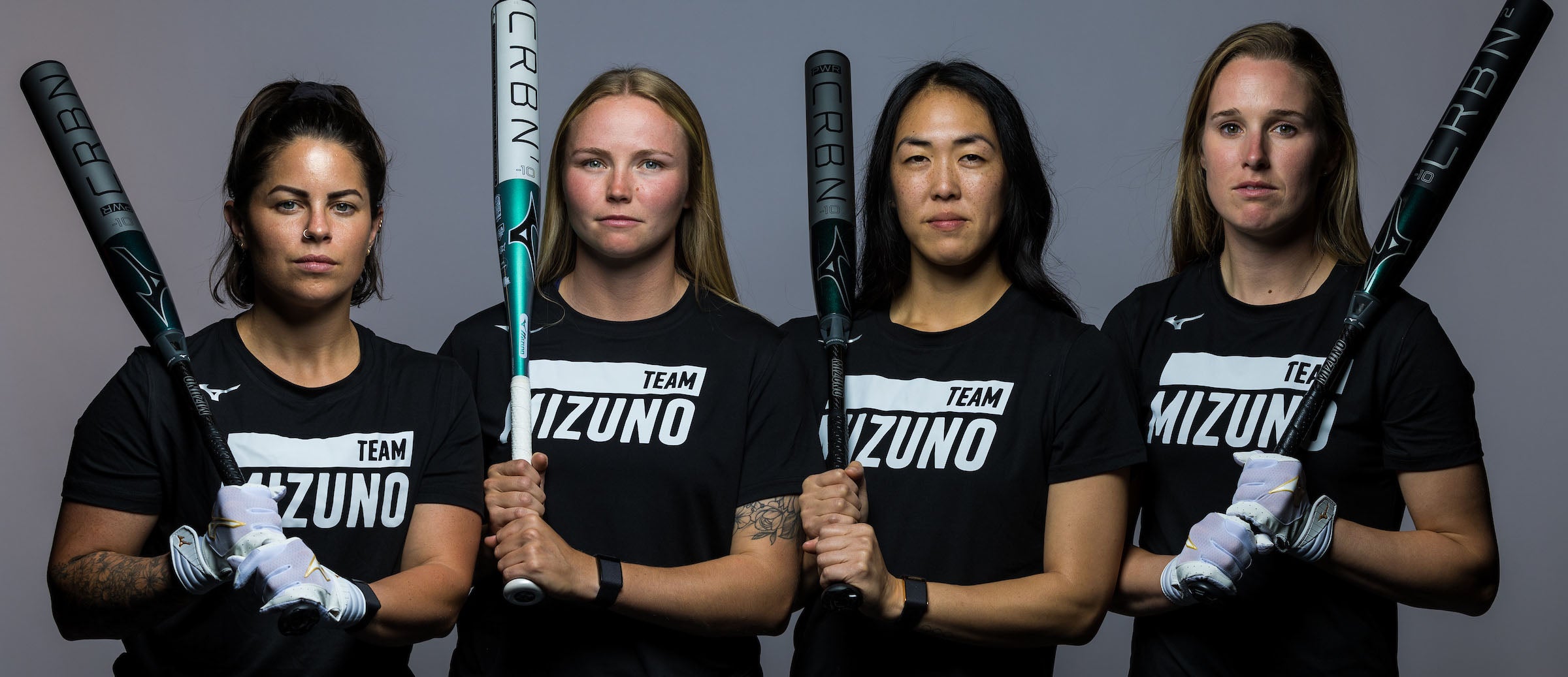 Four women in black Team Mizuno shirts stand side by side holding softball bats vertically, looking confidently at the camera against a plain background.