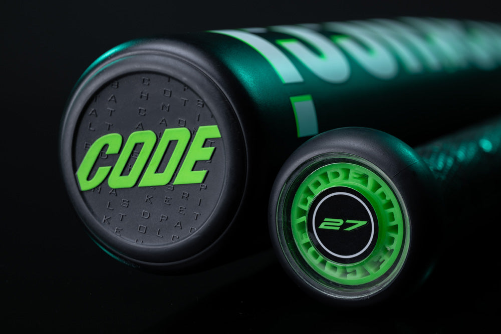 Close-up of two Marucci 2025 Code (-3) BBCOR bats—one displaying "CODE" in bold green with an anti-vibration knob, the other featuring a green circular "27" design.