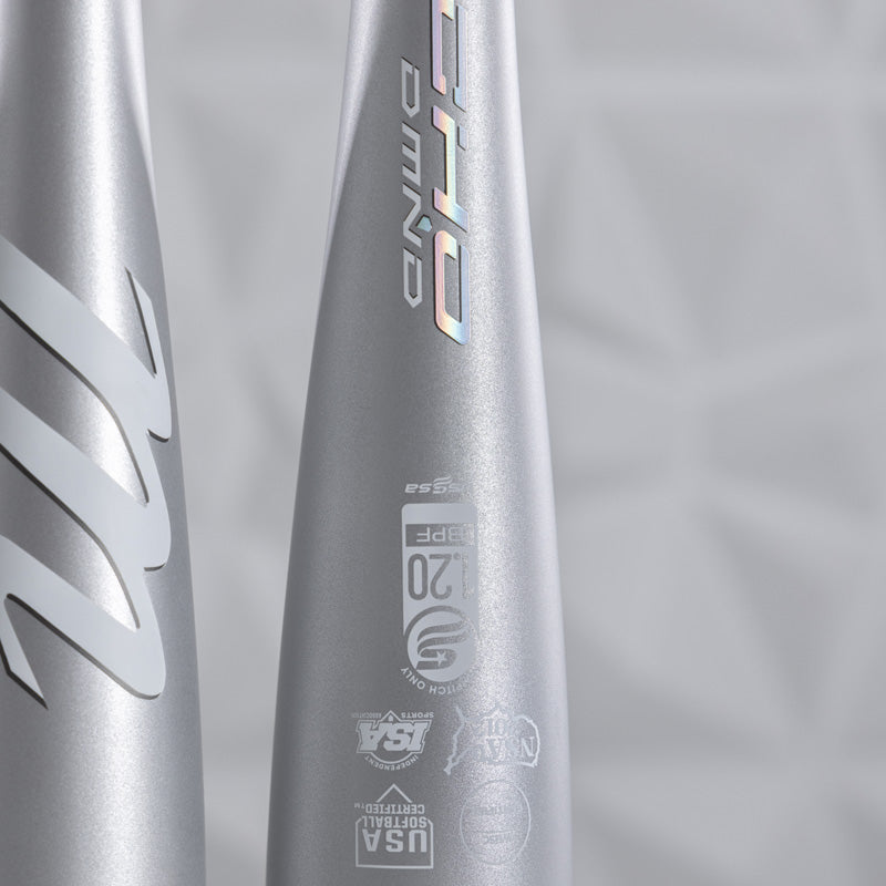 Close-up of two silver baseball bats with certification logos and holographic “D MND” text visible on the barrels, set against a light, geometric background.