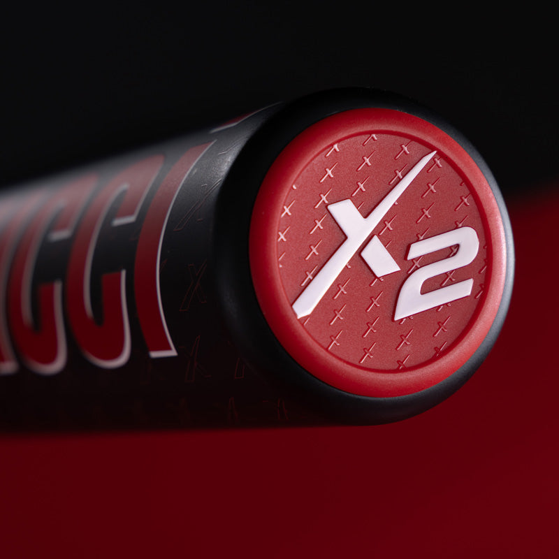 Close-up of the end cap of a black and red baseball bat, featuring a red circle with the white X2 logo embossed on it. The background is dark with hints of red.