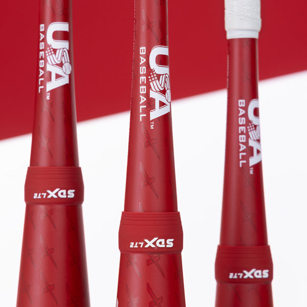 Three red baseball bats with USA Baseball and SLX ZLT printed on them, standing upright against a red and white background. One bat has a white grip at the top.