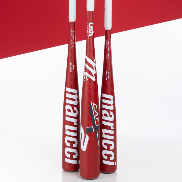 Three red Marucci baseball bats with white handles are standing upright. The Marucci logo and model name are prominently displayed in large white letters on each bat. The background features red and white geometric shapes.