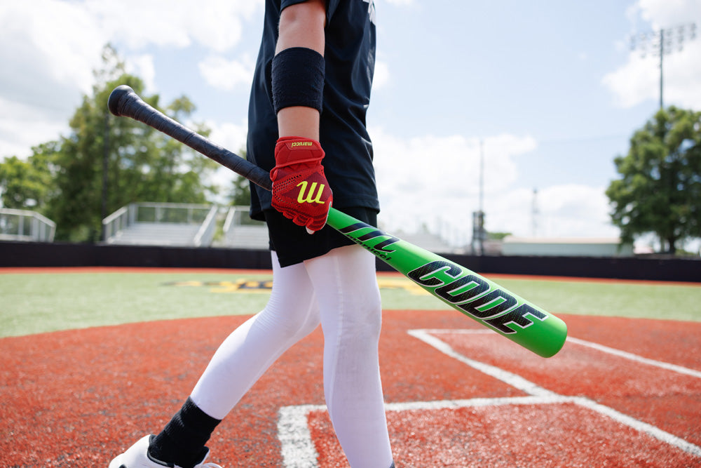 A baseball player in white pants and red gloves holds a green Marucci 2025 Code (-10) USA Baseball Bat (MSBCD10USA) while walking across an orange field with white lines, with bleachers and trees in the background.