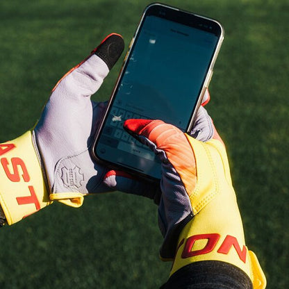 Someone wearing Easton Walk Off Ethos Youth Batting Gloves (WO25YBG) by Easton uses a smartphone outdoors, typing on the touchscreen with their thumb on a grassy field.