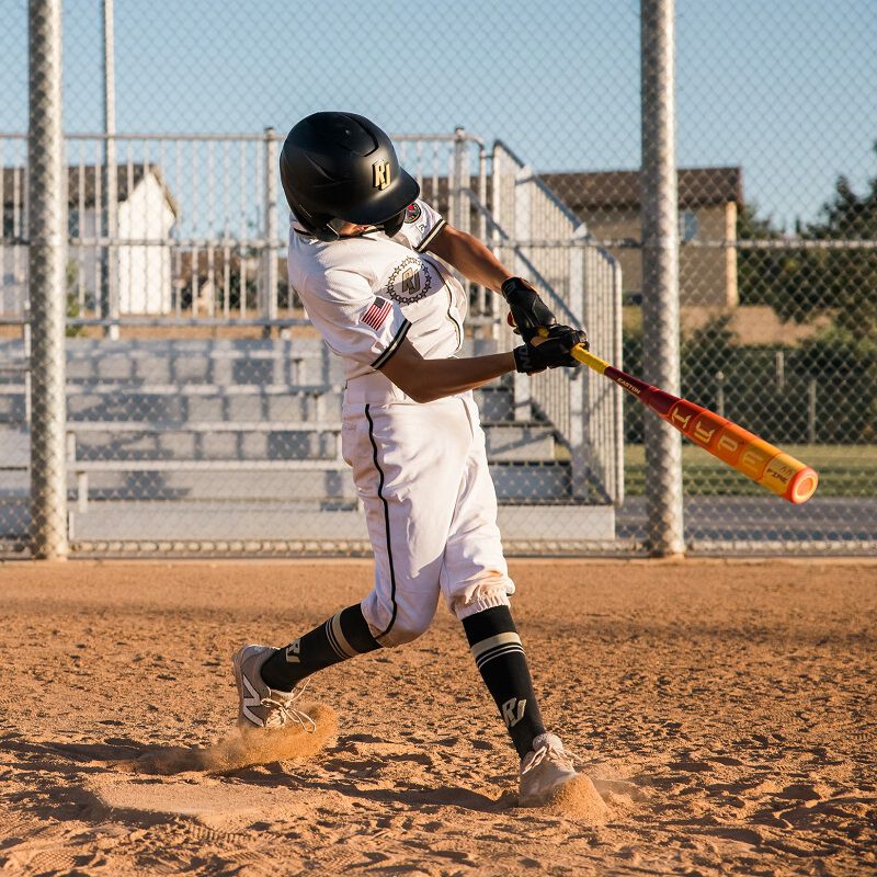 A young baseball player in a white uniform and black helmet swings the 2025 Easton Hype Fire (-5) USSSA Baseball Bat (EUT5HYP5) by Easton on a dirt field, with empty bleachers and a chain-link fence in the background.