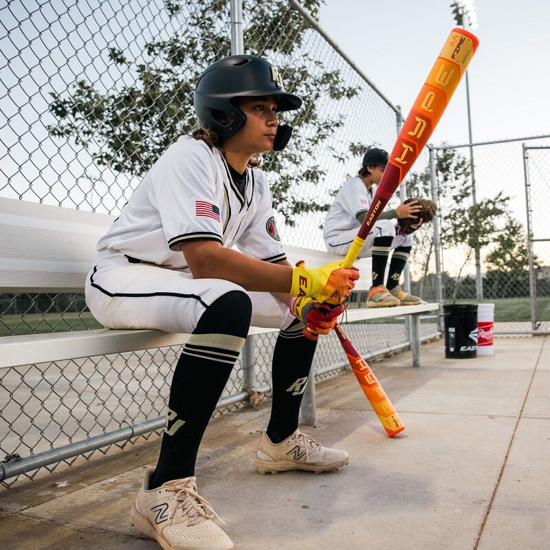 A young baseball player in uniform and helmet sits on a bench holding the 2025 Easton Hype Fire JBB (-10) 2 3/4" USSSA Baseball Bat (EJB5HYP10), with another player and a field visible in the background.