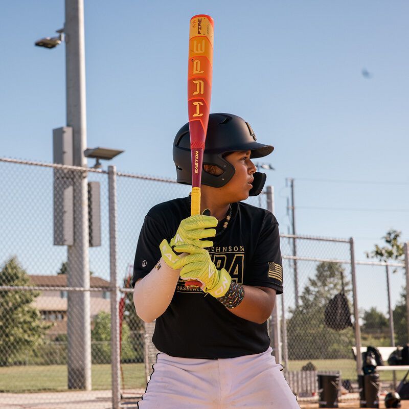 A young baseball player wearing a helmet and gloves stands ready to bat with the 2025 Easton Hype Fire JBB (-10) 2 3/4" USSSA Baseball Bat (EJB5HYP10) on a sunny field, a chain-link fence in the background.