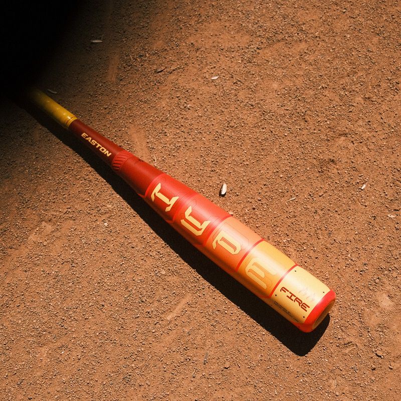 A 2025 Easton Hype Fire JBB (-10) 2 3/4" USSSA Baseball Bat (EJB5HYP10) rests on a dirt field, sunlight highlighting the Easton brand and casting shadows across the textured surface.