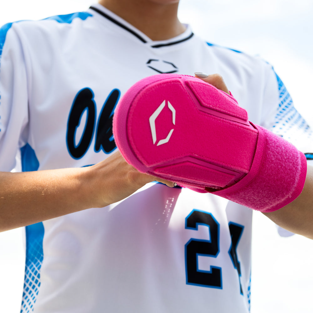A person in a white and blue jersey with the number 21 fastens an EvoShield Sliding Mitt 2.0 (WB57537) by EvoShield, bright pink for added hand protection, on their left hand.
