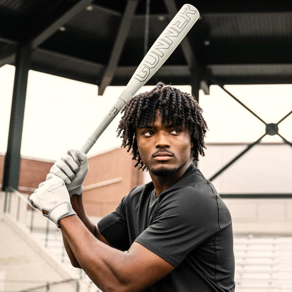 A young man with curly hair, wearing a black shirt and white gloves, holds a baseball bat labeled GUNNER over his shoulder, preparing to swing at an outdoor stadium.