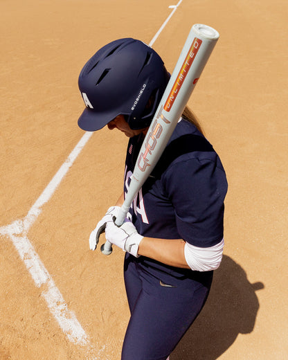 A softball player in a navy uniform and helmet stands on a dirt field near the foul line, holding an Easton 2025 Ghost Unlimited (-10) Fastpitch Softball Bat (EFP5GHUL10) over her shoulder, looking down.