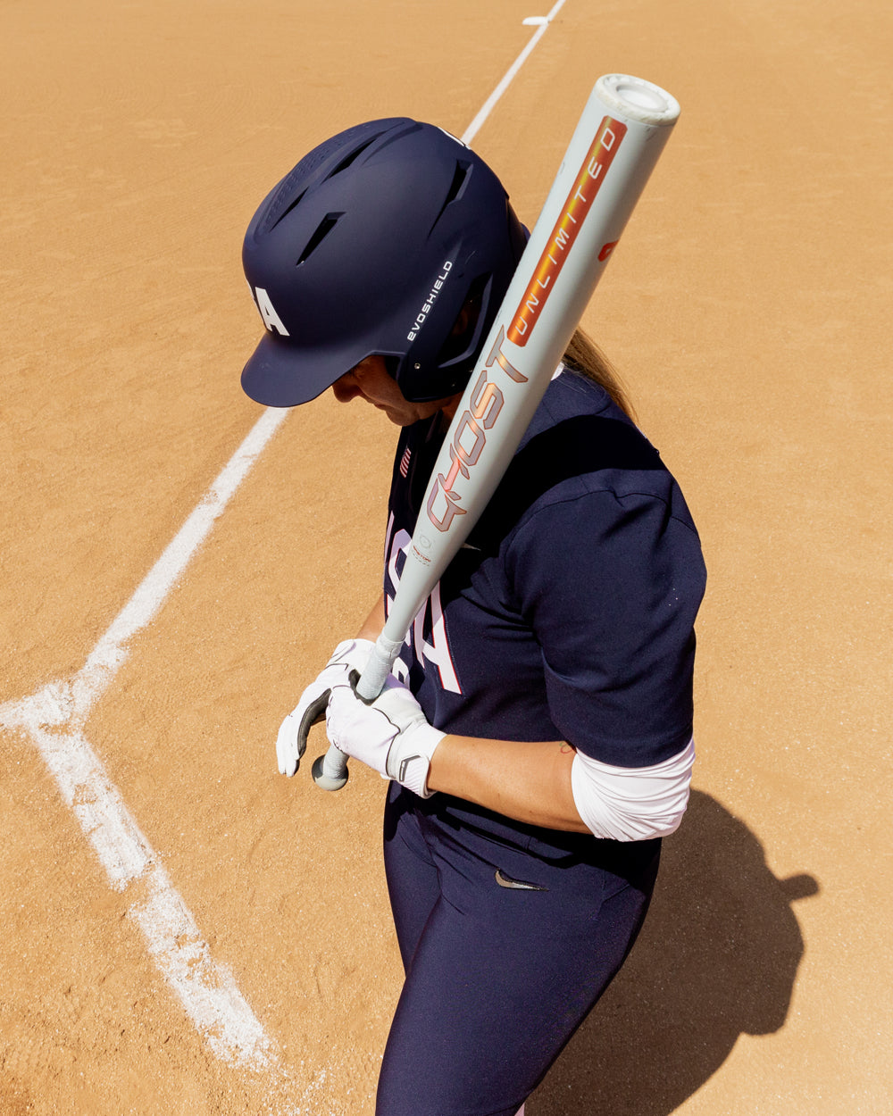 A softball player in a navy uniform and helmet stands by the foul line, holding an Easton 2025 Ghost Unlimited (-9) Fastpitch Softball Bat (EFP5GHUL9) over her shoulder, and looking down.