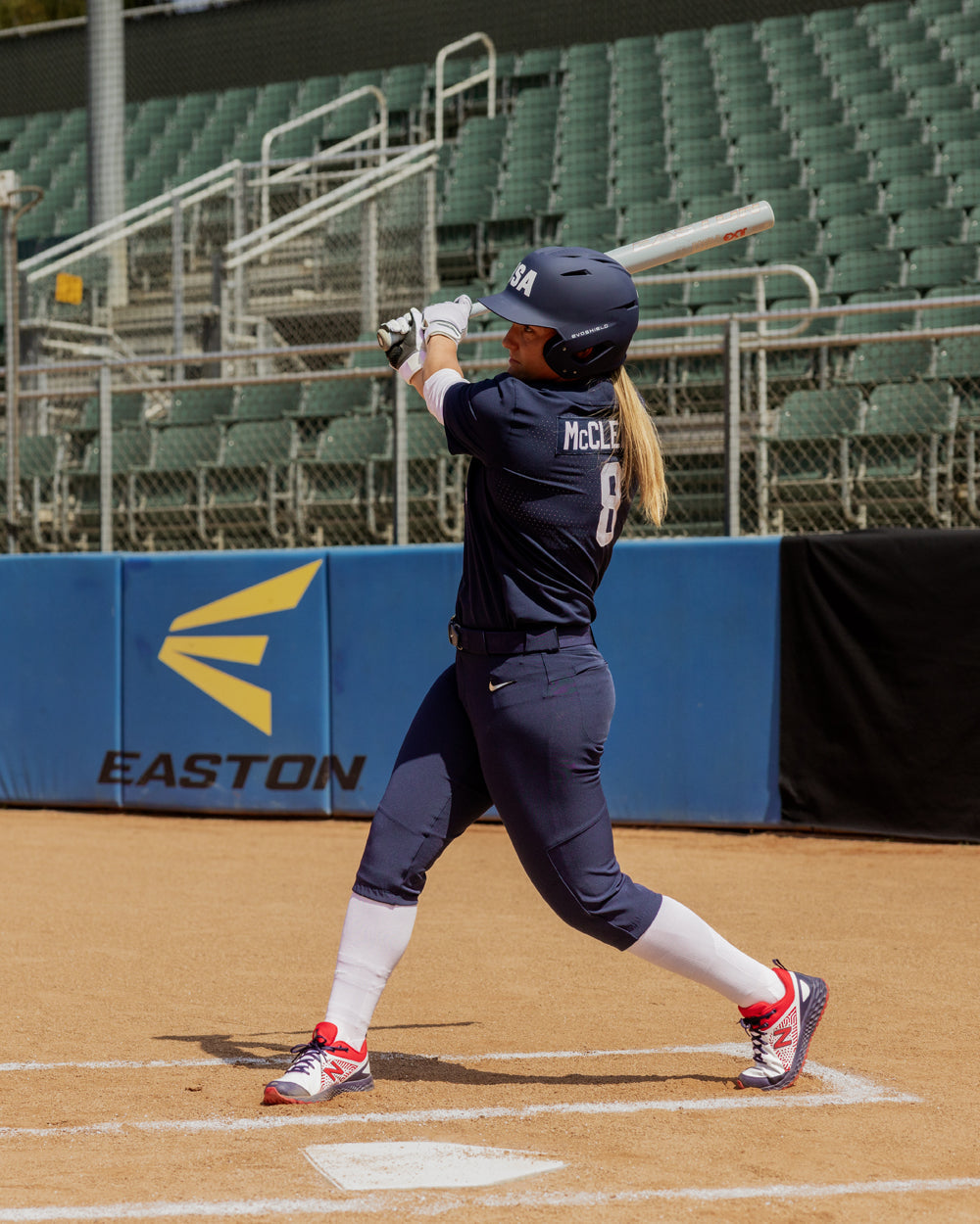 A softball player in a navy blue USA uniform swings the 2025 Easton Ghost Unlimited (-9) Fastpitch Softball Bat (EFP5GHUL9), with empty stadium seats and a blue EASTON banner visible in the background.