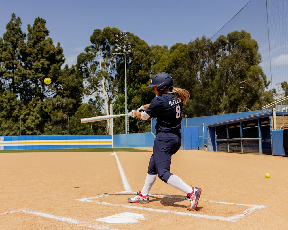 A softball player in navy swings the 2025 Easton Ghost Unlimited (-9) Fastpitch Softball Bat (EFP5GHUL9) by Easton at a ball on a sunny field, with trees and blue fencing in the background.