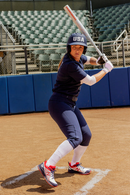 A softball player in a navy USA uniform and helmet stands ready to bat at home plate, gripping her 2025 Easton Ghost Unlimited (-9) Fastpitch Softball Bat (EFP5GHUL9) by Easton. An empty stadium and fence appear in the background of the outdoor field.