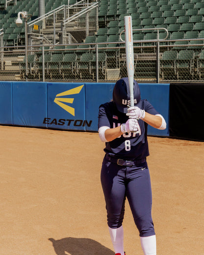 A softball player in a navy blue USA uniform stands on the field, holding a 2025 Easton Ghost Unlimited (-9) Fastpitch Softball Bat (EFP5GHUL9) vertically in front of her face. The empty stands and Easton logo are visible behind her.