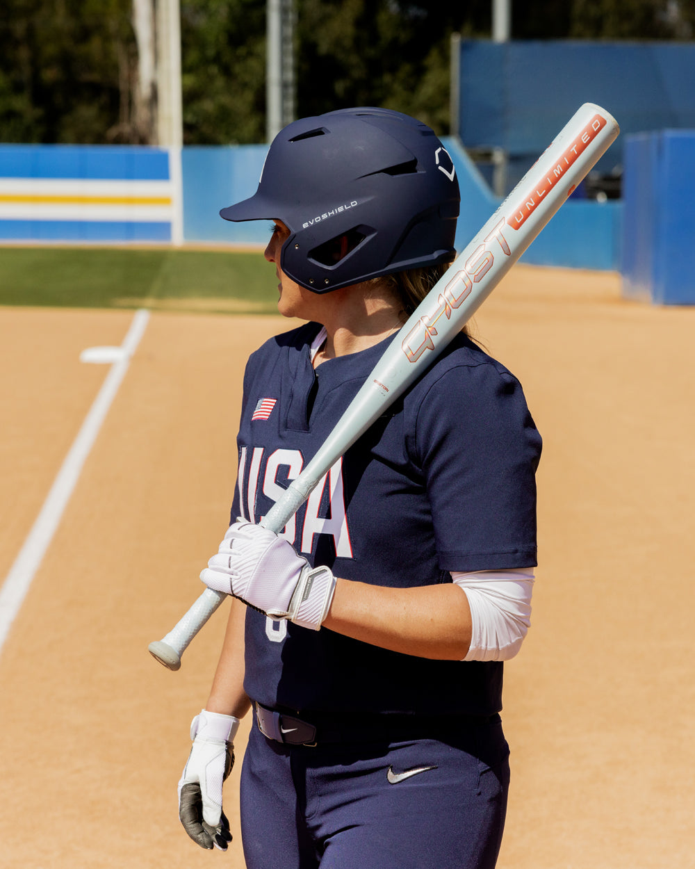 A softball player in a navy blue USA uniform stands on the field, holding the 2025 Easton Ghost Unlimited (-11) Fastpitch Softball Bat (DEMO) by Easton over her shoulder. A dirt infield and blue and yellow fence appear in the background.