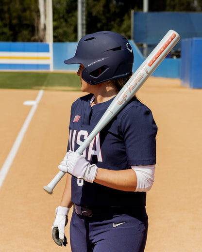 A softball player in a navy blue USA uniform stands on the field, holding the 2025 Easton Ghost Unlimited (-11) Fastpitch Softball Bat (DEMO) by Easton over her shoulder. A dirt infield and blue and yellow fence appear in the background.