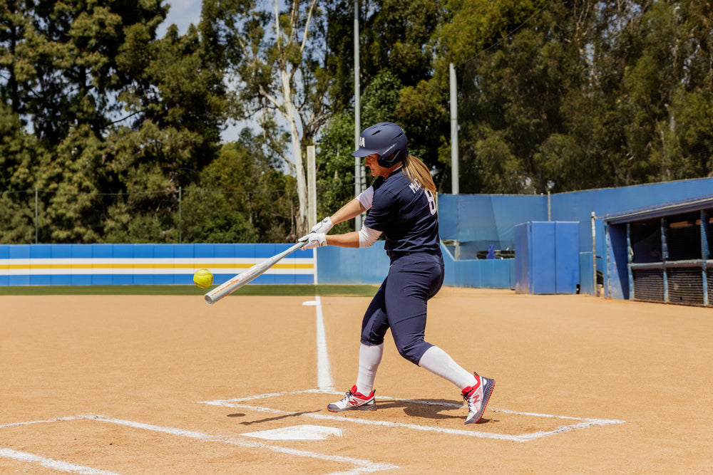 A softball player in a navy uniform swings a 2025 Easton Ghost Unlimited (-9) Fastpitch Softball Bat (EFP5GHUL9) by Easton at a yellow softball on a sunny field, with trees and a blue dugout visible in the background.