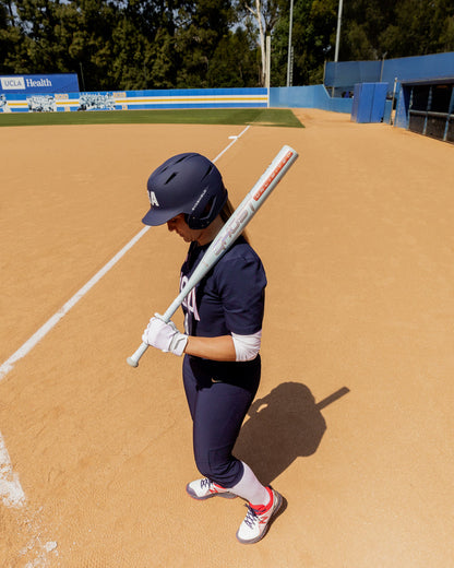 A softball player in a navy blue uniform and helmet holds the 2025 Easton Ghost Unlimited (-9) Fastpitch Softball Bat (EFP5GHUL9) by Easton over their shoulder on a sandy field near home plate, with dugouts and trees visible in the background.