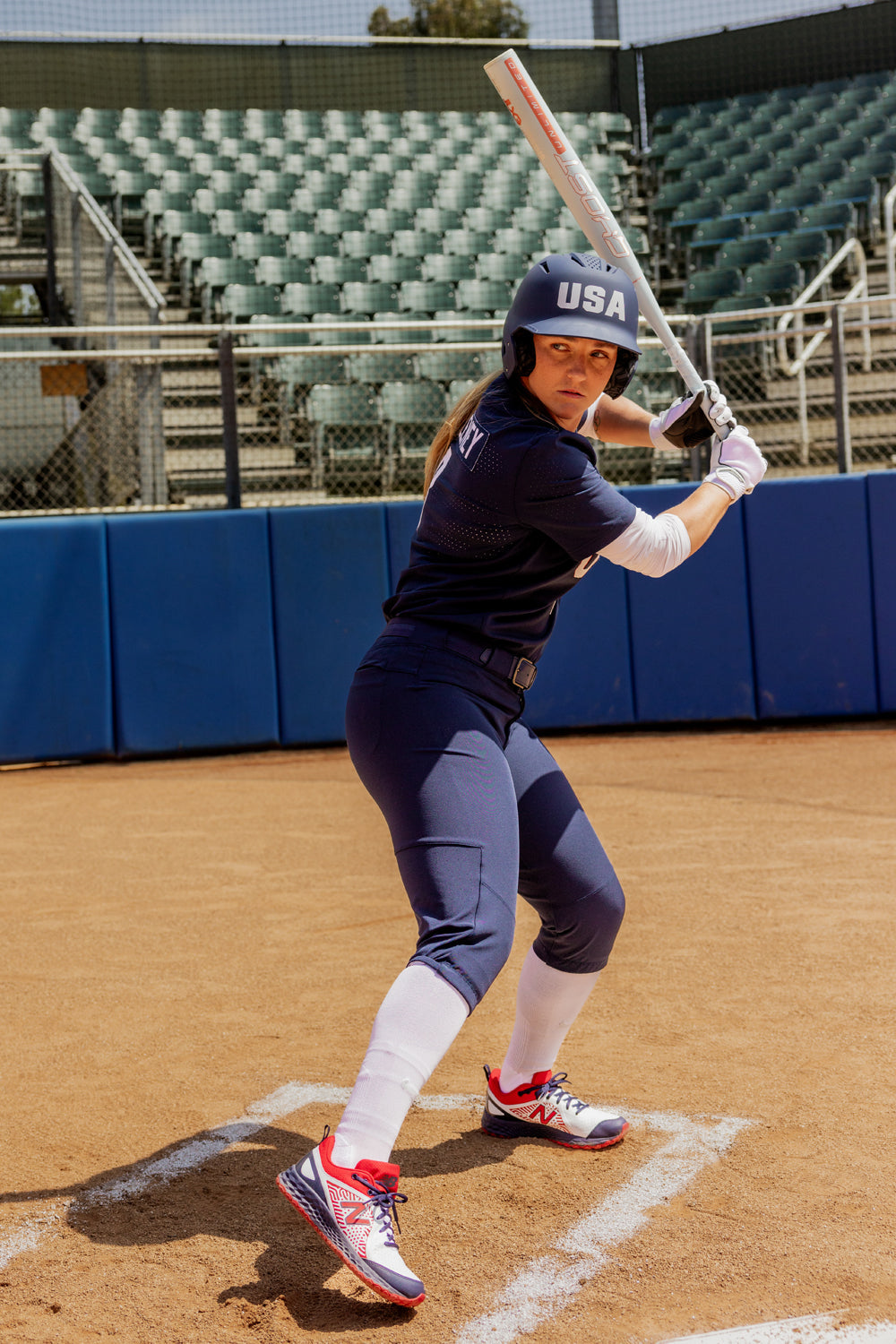 A softball player in a navy USA uniform stands at home plate with the 2025 Easton Ghost Unlimited (-9) Fastpitch Softball Bat (EFP5GHUL9), ready to hit, as empty green bleachers fill the background.