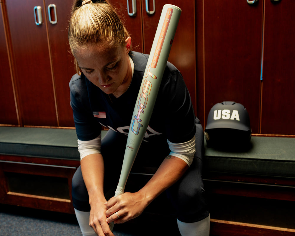 A softball player in a USA uniform sits on a locker room bench, holding her 2025 Easton Ghost Unlimited (-11) Fastpitch Softball Bat (EFP5GHUL11 DEMO) upright, with a USA helmet beside her.