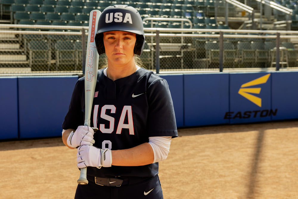 A softball player in a navy USA uniform and helmet stands on a dirt field holding the 2025 Easton Ghost Unlimited (-11) Fastpitch Softball Bat (EFP5GHUL11 DEMO), with empty bleachers and an Easton sign visible in the background.