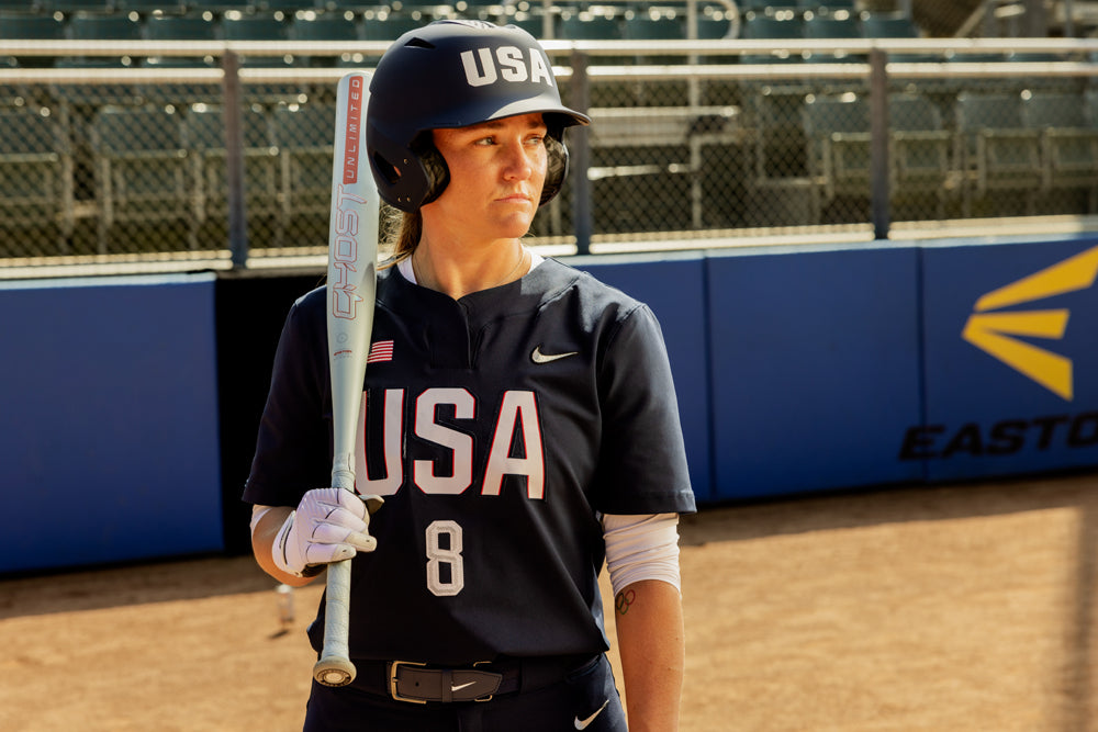A softball player in a navy USA uniform and helmet stands on the field holding an Easton 2025 Ghost Unlimited (-9) Fastpitch Softball Bat (EFP5GHUL9), number 8 visible on her jersey, with empty stadium seats in the background.