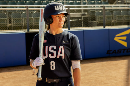 A softball player in a navy USA uniform and helmet stands on the field holding an Easton 2025 Ghost Unlimited (-9) Fastpitch Softball Bat (EFP5GHUL9), number 8 visible on her jersey, with empty stadium seats in the background.