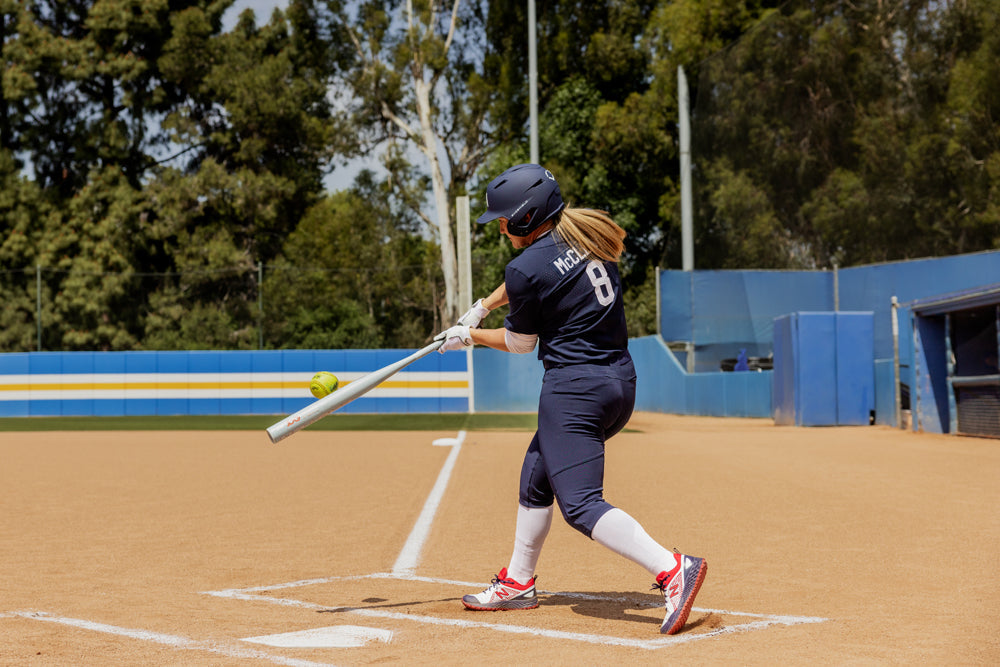 A softball player in a navy uniform swings the 2025 Easton Ghost Unlimited (-9) Fastpitch Softball Bat (EFP5GHUL9) and hits a green ball on an outdoor field. The player’s jersey shows number 8, with trees and a blue fence behind them.