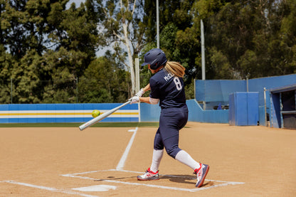 A softball player in a navy uniform swings the 2025 Easton Ghost Unlimited (-9) Fastpitch Softball Bat (EFP5GHUL9) and hits a green ball on an outdoor field. The player’s jersey shows number 8, with trees and a blue fence behind them.
