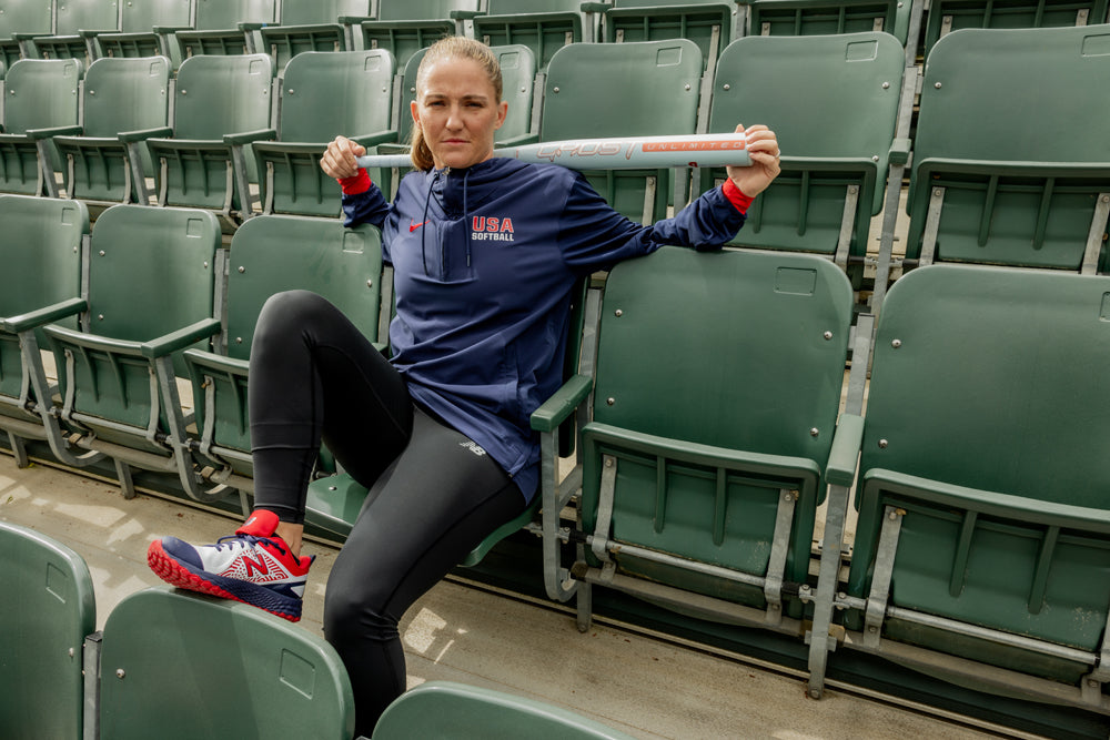 A woman wearing a USA Softball jacket sits in empty stadium seats, holding the 2025 Easton Ghost Unlimited (-10) Fastpitch Softball Bat (EFP5GHUL10) across her shoulders as she looks toward the camera, dressed in athletic shoes and black leggings.
